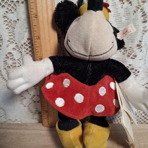 Pu&ograve; includere: Una bambola di peluche di Minnie Mouse con corpo nero, guanti bianchi e scarpe gialle. La bambola indossa una gonna rossa a pois bianchi e ha una faccia sorridente. Un righello di legno &egrave; accanto alla bambola.