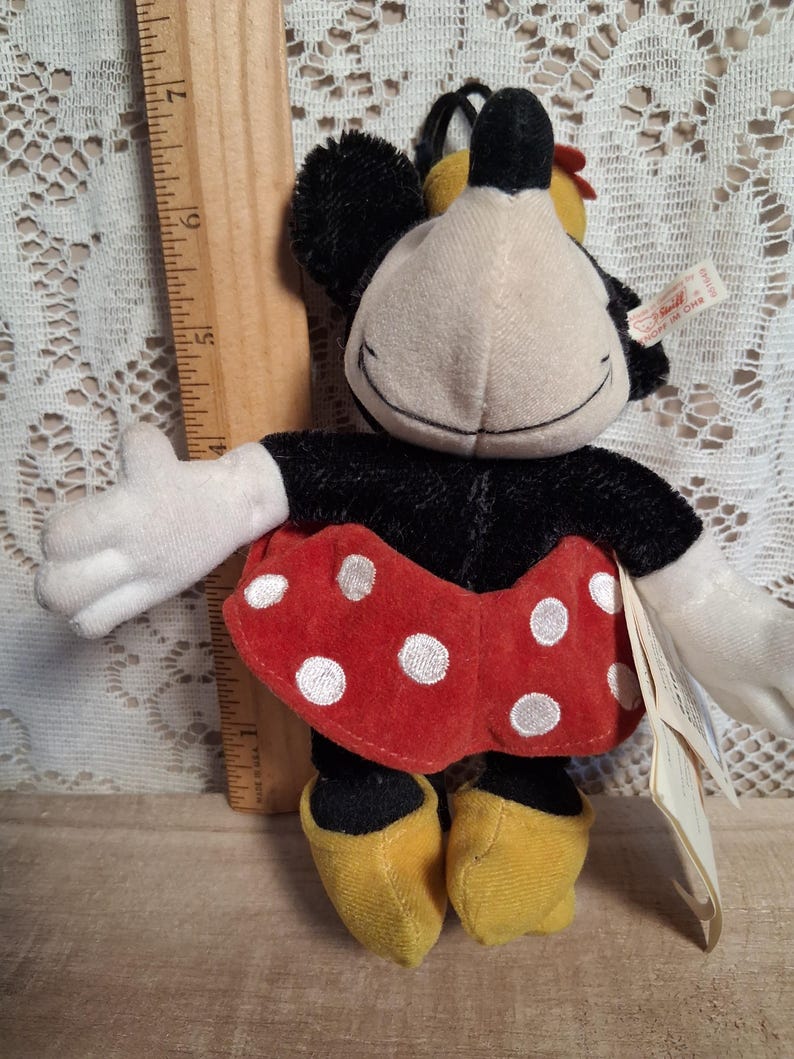Pu&ograve; includere: Una bambola di peluche di Minnie Mouse con corpo nero, guanti bianchi e scarpe gialle. La bambola indossa una gonna rossa a pois bianchi e ha una faccia sorridente. Un righello di legno &egrave; accanto alla bambola.