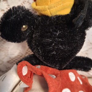 Pu&ograve; includere: Un giocattolo di peluche nero vintage con un cappello giallo e pantaloncini a pois rossi. Il giocattolo ha un occhio a bottone e guanti bianchi. Il peluche &egrave; un oggetto da collezione.