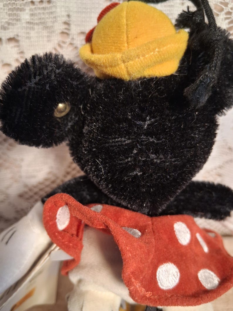 Pu&ograve; includere: Un giocattolo di peluche nero vintage con un cappello giallo e pantaloncini a pois rossi. Il giocattolo ha un occhio a bottone e guanti bianchi. Il peluche &egrave; un oggetto da collezione.