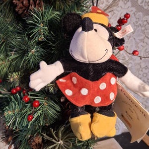 Pu&ograve; includere: Ornamento in peluche di Minnie Mouse con gonna rossa a pois bianchi, pelliccia nera e scarpe gialle. L'ornamento ha un cappello giallo e guanti bianchi. &Egrave; appeso a un albero di Natale con bacche rosse e pigne.