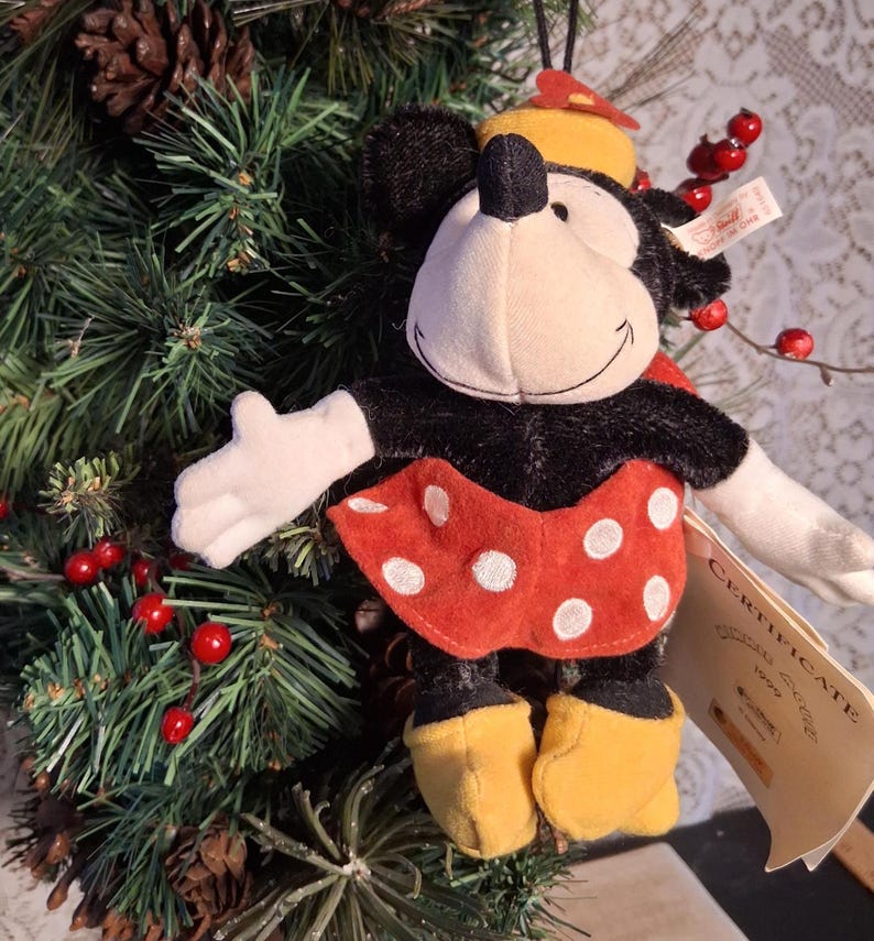 Pu&ograve; includere: Ornamento in peluche di Minnie Mouse con gonna rossa a pois bianchi, pelliccia nera e scarpe gialle. L'ornamento ha un cappello giallo e guanti bianchi. &Egrave; appeso a un albero di Natale con bacche rosse e pigne.