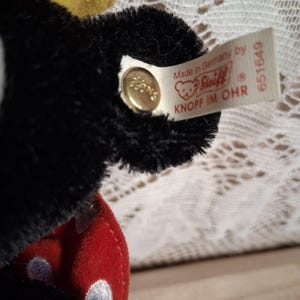 Pu&ograve; includere: Primo piano di un peluche nero con un design a pois rossi e bianchi. Il giocattolo ha un accento giallo e un'etichetta che dice "Made in Germany by Steiff". Un bottone dorato &egrave; attaccato all'orecchio.