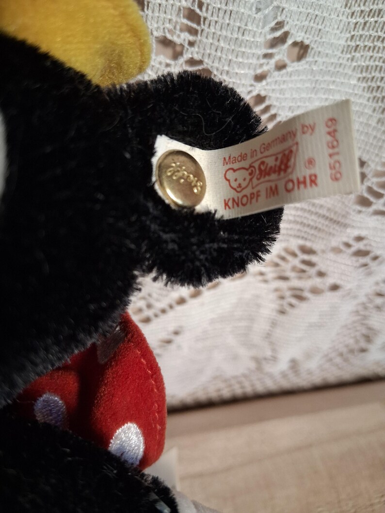 Pu&ograve; includere: Primo piano di un peluche nero con un design a pois rossi e bianchi. Il giocattolo ha un accento giallo e un'etichetta che dice "Made in Germany by Steiff". Un bottone dorato &egrave; attaccato all'orecchio.