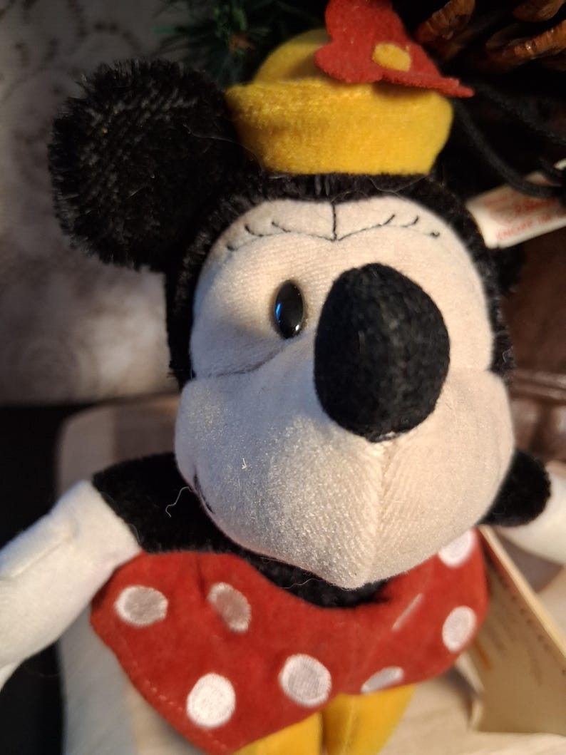 Pu&ograve; includere: Una bambola di peluche di Minnie Mouse con testa e orecchie nere, viso bianco e fiocco a pois rossi e bianchi. Indossa un cappello giallo con un fiore rosso. La bambola ha un colletto a pois rossi e bianchi e scarpe gialle.