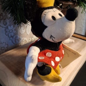 Pu&ograve; includere: Bambola di peluche di Minnie Mouse, con un vestito rosso a pois bianchi, scarpe gialle e un cappello giallo con un fiocco rosso. La bambola ha orecchie nere e un naso nero. &Egrave; attaccata una piccola etichetta.
