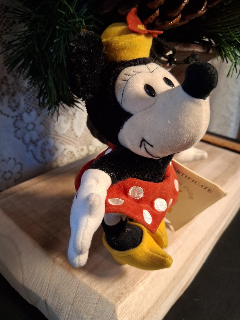 Pu&ograve; includere: Bambola di peluche di Minnie Mouse, con un vestito rosso a pois bianchi, scarpe gialle e un cappello giallo con un fiocco rosso. La bambola ha orecchie nere e un naso nero. &Egrave; attaccata una piccola etichetta.