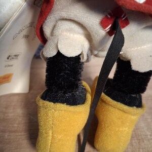 Pu&ograve; includere: Primo piano di un peluche di Minnie Mouse. Il giocattolo ha gambe nere e pelose e scarpe gialle. L'outfit include un vestito bianco con orlo smerlato e un fiocco rosso. &Egrave; visibile un fiocco rosso a pois bianchi.