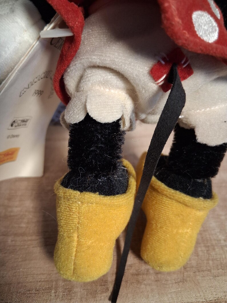 Pu&ograve; includere: Primo piano di un peluche di Minnie Mouse. Il giocattolo ha gambe nere e pelose e scarpe gialle. L'outfit include un vestito bianco con orlo smerlato e un fiocco rosso. &Egrave; visibile un fiocco rosso a pois bianchi.
