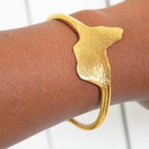 May include: A gold-colored cuff bracelet with a textured, abstract design. The bracelet is worn on a person's arm. The cuff is made of a shiny metal and has a unique, organic shape. The bracelet is a stylish accessory.