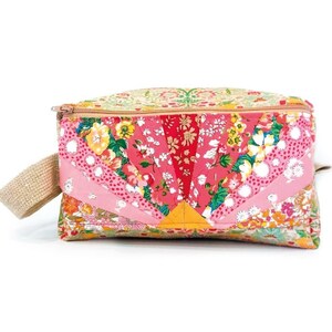 Puede incluir: Una bolsa de cremallera con estampado floral rosa y amarillo con un asa de tela beige. La bolsa está hecha de tela de patchwork con una variedad de estampados florales.
