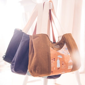 May include: Three tote bags hanging on a white rack. The bag on the right is brown with a floral pattern and a light brown handle. The bag in the middle is dark blue with a light brown handle. The bag on the left is dark blue with a black handle.