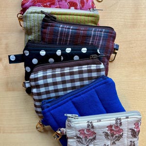 May include: A selection of small zipped pouches in diverse patterns and colours. Designs include floral, polka dot, plaid, and quilted textures. Each pouch has a metal zip and a small loop for attaching to a keyring.