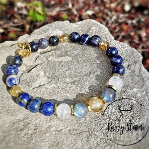 May include: A gemstone bracelet with blue, gold, and white beads. The bracelet is presented on a grey stone surface, with a blurred natural backdrop. The bracelet incorporates various round beads and gold-coloured spacers.