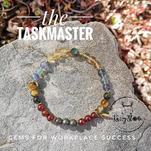 May include: A multi-colored gemstone bracelet with gold beads and a gold charm that says "Karzy Stone". The bracelet is on a gray rock with the text "the TASKMASTER" above it and "GEMS FOR WORKPLACE SUCCESS" below it.