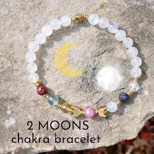 Peut inclure: Bracelet en pierres de lune blanches avec des breloques en forme de croissant de lune dorées et des pierres de chakra colorées. Le bracelet est sur un rocher gris avec le texte "2 MOONS chakra bracelet".
