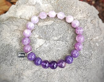 Puede incluir: Un brazalete de piedras preciosas de color púrpura y lavanda con un dije de plata que dice "Moxi". El brazalete está hecho de cuentas redondas y está sobre un fondo de roca gris.