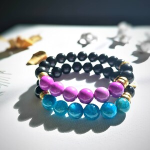 May include: Two bracelets made of black, pink, and blue beads. The top bracelet is made of black beads with gold accents. The bottom bracelet is made of pink and blue beads.