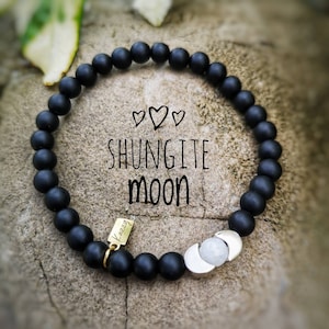 Può includere: Braccialetto di perline nere con un ciondolo a forma di luna argentato e un'etichetta dorata con la scritta "Kazzy". Il braccialetto è su uno sfondo marrone con le parole "Shungite Moon" scritte in bianco.