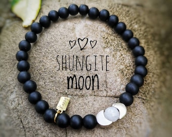 May include: Black beaded bracelet with a silver moon charm and a gold tag that says "Kazzy". The bracelet is on a brown background with the words "Shungite Moon" written in white.