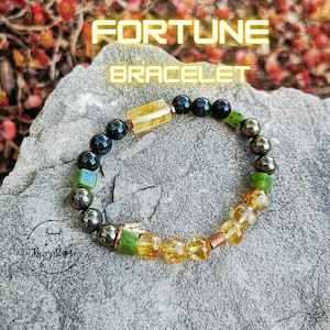 Puede incluir: Un brazalete de cuentas doradas y verdes con el texto "FORTUNE BRACELET" sobre un fondo de roca gris.