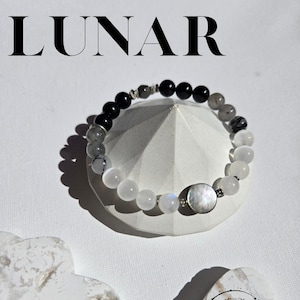 Peut inclure: Un bracelet de perles avec un mélange de pierres noires, grises et blanches, dont une grande pièce centrale ronde et irisée. Le mot "LUNAR" est affiché en grandes lettres noires en haut de l'image. Le bracelet est présenté sur une forme géométrique blanche.