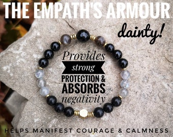 May include: Black and grey beaded bracelet with gold accents. The bracelet has a hexagonal charm and the text "Provides strong protection & absorbs negativity" is printed on a white background.