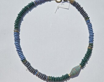 Puede incluir: Un collar artesanal con una mezcla de cuentas en forma de disco azules, verdes y grises. El collar incluye una cuenta central alargada con un patrón de rayas y detalles dorados. El cierre es de color dorado.