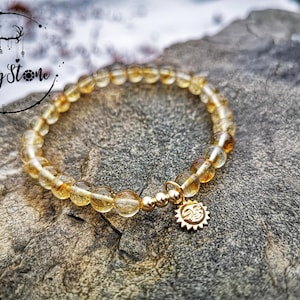 Puede incluir: Un brazalete dorado con un dije de sol hecho de cuentas de citrino amarillo.