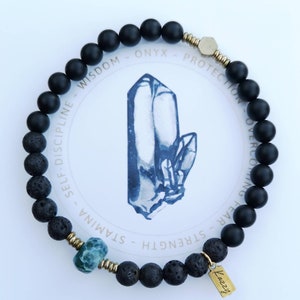 May include: Black onyx and lava rock beaded bracelet with gold accents and a blue gemstone. The bracelet has the words "Wisdom - Onyx - Protection - Overcoming Fear - Strength - Stamina - Self-Discipline" written around the beads.