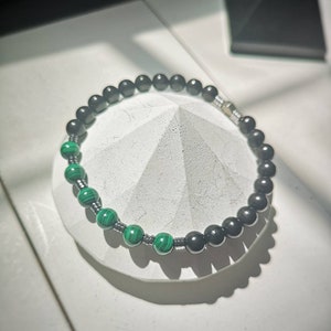 May include: A beaded bracelet featuring a combination of black and green beads. The bracelet has a section of vibrant green malachite beads and a section of black beads. The beads are strung together with a silver-colored metal. The bracelet is displayed on a white surface.