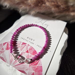 Op de afbeelding: Een delicate armband gemaakt van kleine, gefacetteerde, dieproze robijnkralen met een zilverkleurige metalen sluiting. De armband wordt getoond op een witte kaart met de tekst July, Ruby, Passion, Protection, Prosperity en een edelsteenillustratie.