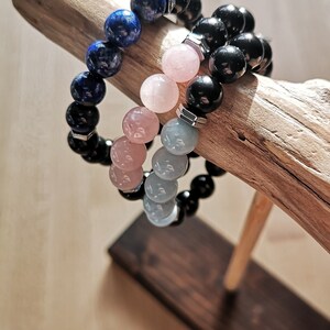 May include: Three beaded bracelets stacked on a wooden stand. The top bracelet is made of dark blue beads, the middle bracelet is made of light pink beads, and the bottom bracelet is made of light gray beads.