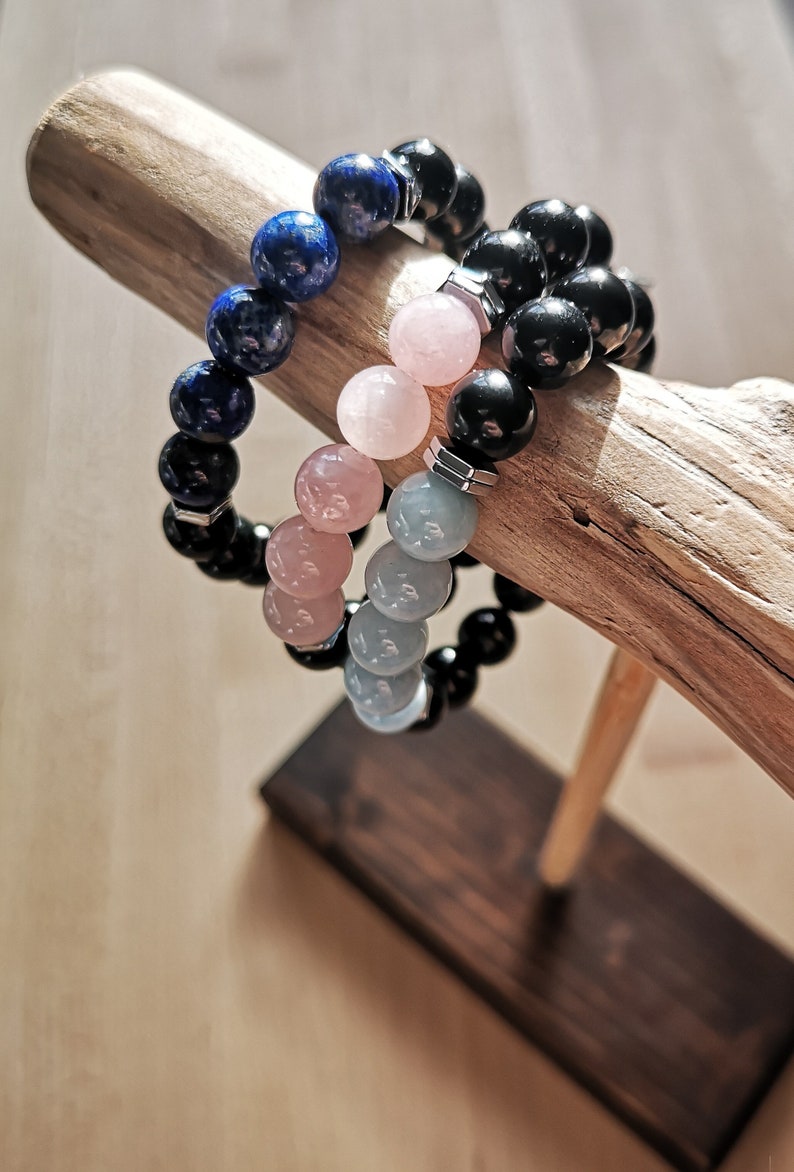 May include: Three beaded bracelets stacked on a wooden stand. The top bracelet is made of dark blue beads, the middle bracelet is made of light pink beads, and the bottom bracelet is made of light gray beads.