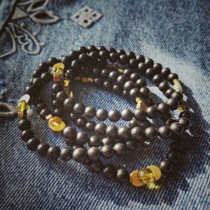 Puede incluir: Un conjunto de pulseras de cuentas negras con detalles dorados. Las pulseras están superpuestas y tienen un acento de piedra preciosa amarilla.