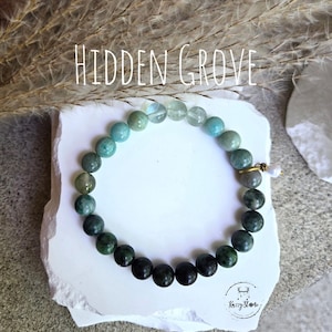 May include: A beaded bracelet featuring a gradient of round stones transitioning from translucent light green to opaque dark forest green. The bracelet includes a small gold-toned metal charm and is labelled with the text HIDDEN GROVE.