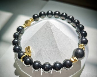 Puede incluir: Un brazalete de cuentas de hematites negras con detalles dorados y una pequeña etiqueta dorada que dice "W.M."