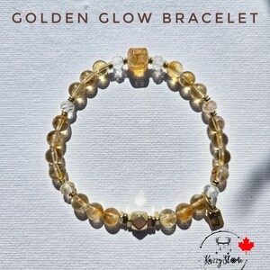 May include: A golden glow bracelet with a mix of light and dark yellow beads. The bracelet has a gold clasp and a small tag with the text "Kizzy Stone" on it.