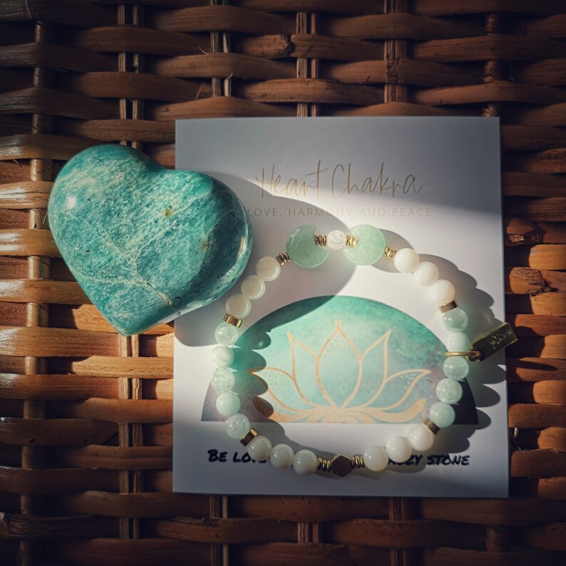 May include: A green heart-shaped stone with a green and white bracelet with gold accents. The bracelet has the words "Heart Chakra" and "Love, Harmony and Peace" on the card. The card also has a green lotus flower design and the words "Be Love" and "Aley Stone".