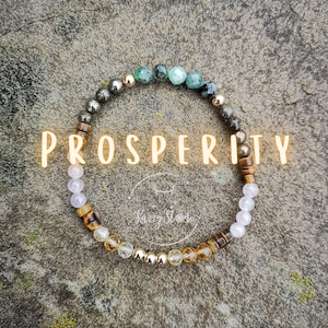 May include: A bracelet made of various gemstones, including green, brown, gold, and white beads. The bracelet is on a gray, textured surface. The word "PROSPERITY" is written in yellow letters on the image.