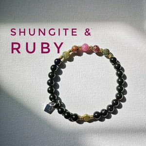 Könnte beinhalten: Ein schwarzes Perlenarmband mit goldenen Akzenten und einem einzigen rosafarbenen und grünen Edelstein. Das Armband trägt die Aufschrift "Shungite & Ruby".