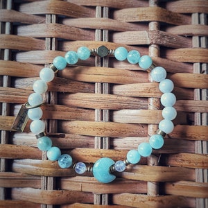May include: A light blue and white beaded bracelet with a crescent moon charm. The bracelet is made with aquamarine and moonstone beads and has a gold clasp.