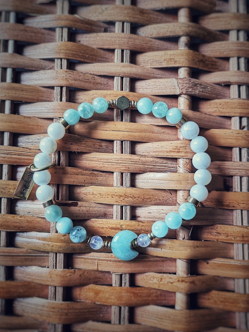 May include: A light blue and white beaded bracelet with a crescent moon charm. The bracelet is made with aquamarine and moonstone beads and has a gold clasp.