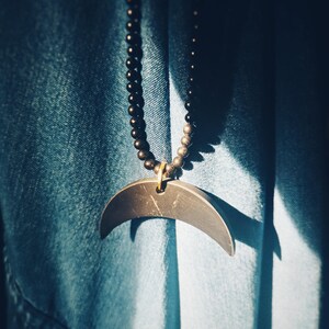 Può includere: Una collana di perline nere con un ciondolo a forma di mezzaluna in metallo scuro. La collana è appesa a un tessuto di jeans blu.