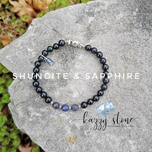 Può includere: Braccialetto di perline nere con chiusura argentata e tre perline di zaffiro blu. Il braccialetto è su una roccia grigia con il testo "Shungite & Sapphire" e "kazzy stone Handmade in Canada".