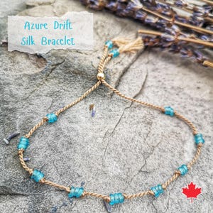 May include: A light brown silk cord bracelet with a gold clasp and blue glass beads. The bracelet is called "Azure Drift Silk Bracelet".
