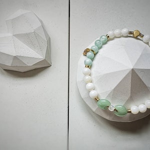 May include: A white and green beaded bracelet with a gold accent. The bracelet is on a white heart-shaped dish.