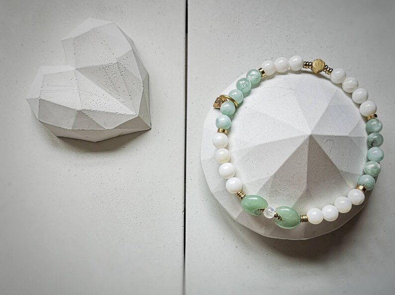 May include: A white and green beaded bracelet with a gold accent. The bracelet is on a white heart-shaped dish.