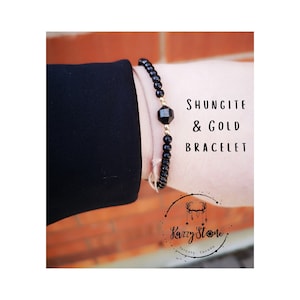 Può includere: Un braccialetto nero in shungite e oro con un ciondolo di pietra nera. Il braccialetto è al polso di una persona. Il testo "Shungite & Gold Bracelet" è scritto sull'immagine.