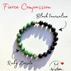 May include: A green and black beaded bracelet with gold accents. The bracelet is made with ruby zoisite and black tourmaline stones. The bracelet is labelled "Fierce Compassion".